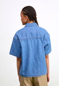 Short-sleeve denim shirt, light blue, relaxed fit, visible stitching details, and a simple collar, worn with beige pants.