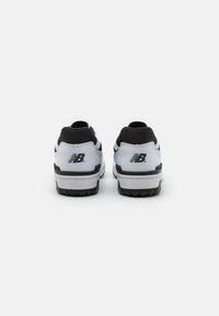 Pari valkoisia ja mustia New Balance -lenkkareita näkyy takaa, jossa on NB-logo kantapäässä, valkoista taustaa vasten.