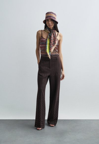 Γυναίκα που φοράει καφέ παντελόνι με σχέδιο ζιγκ-ζαγκ, ριγέ αμάνικη μπλούζα, πολύχρωμο φουλάρι και καπέλο τύπου bucket στο ίδιο χρώμα, στέκεται μπροστά σε απλό φόντο.