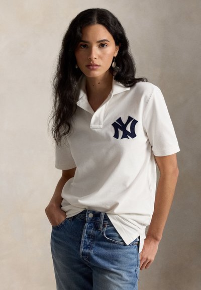 Νέα γυναίκα με μακριά σκούρα μαλλιά, φοράει άσπρο πόλο πουκάμισο με σκούρο λογότυπο και μπλε τζιν, όρθια με το ένα χέρι στην τσέπη.