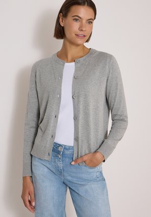 Femme portant un cardigan gris boutonné par-dessus un haut blanc avec un jean bleu clair, debout avec une main dans la poche devant un fond uni.