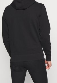 Czarna bluza z kapturem wykonana z gładkiego materiału, wyposażona w duży kaptur, ribbowe mankiety oraz dół. Tył ma czysty design z minimalną ilością szwów.