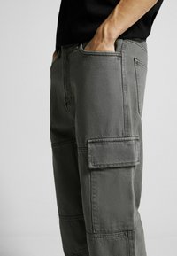 Šedé cargo kalhoty vyrobené z odolné látky, které mají několik kapes, uvolněný střih a viditelné detaily šití.