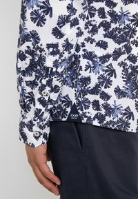 Camicia floreale bianca e blu navy con un design a motivi, dotata di bottoni sui polsini e un'etichetta del marchio visibile all'orlo.