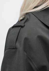 Close-up van het schoudergebied op een steenkoolgrijze jas met een knoopstrookdetail en lichtblond haar zichtbaar.