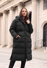 Abrigo de plumas negro largo con capucha, que presenta un patrón acolchado y botones en el frente. Dos bolsillos laterales y un dobladillo ligeramente acampanado.