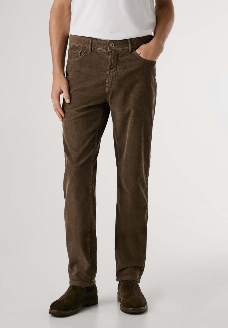 Homme portant un pantalon en velours côtelé marron, des bottes en daim marron foncé et une chemise blanche, debout avec une main dans la poche sur un fond uni.