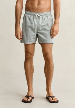 Man draagt lichtgrijze zwembroek met witte koord en bruine teenslippers, staand tegen een beige achtergrond.