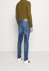 Kapeuden farkut siniset denim housut, joissa on vaalea kuluminen, ruskea nahkasauma takana, yhdistettynä valkoisiin tennareihin ja vihreään pitkähihasta paita.