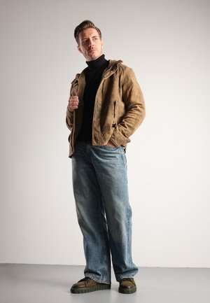 Hnědá suede bunda s kapucí, černý rolák, světle modré džíny a boty se zelenou podrážkou. Oblečení má volný střih a ležérní styl.