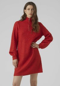 Abito rosso in maglia con colletto alto, texture diagonale, maniche lunghe a sbuffo e dettaglio con zip a quarto. Silhouette liscia e aderente.