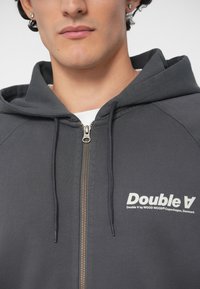 Σκούρο γκρι φούτερ με φερμουάρ, κατασκευασμένο από μαλακό ύφασμα, με φερμουάρ στο μπροστινό μέρος, κορδόνια και λευκό λογότυπο στην αριστερή περιοχή του στήθους.