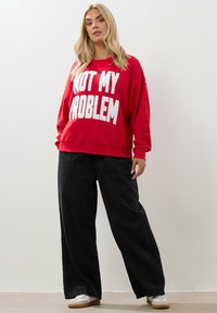 Felpa rossa con testo bianco "NON È UN MIO PROBLEMA", vestibilità ampia, polsini a costine, abbinata a pantaloni neri a gamba larga e sneakers bianche.
