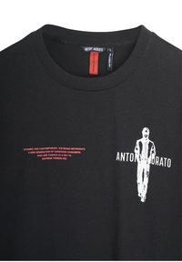 Crna pamučna majica s bijelom grafikom figure i crvenim tekstom sprijeda. Oznaka u izrezu označava naziv branda.