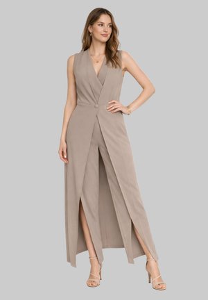 Kobieta ubrana w bezrękawnikowy kombinezon typu kopertowego w kolorze taupe z przednimi rozcięciami oraz beżowe sandały na wysokim obcasie na cienkich paskach, pozująca z ręką na biodrze.