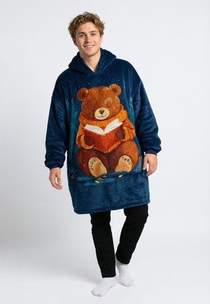 Oversized marineblauw pluche hooded deken met een berenillustratie die een boek leest. Heeft een zachte textuur en lange mouwen. Er worden zwarte broek onder gedragen.