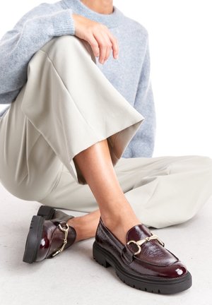 Bordeaux leren instappers met een opvallende gouden gespsluiting en een dikke zwarte rubberen zool. De outfit bevat lichtgrijze wijd-uitlopende broek.