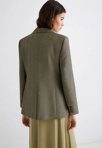 Blazer verde oliva con un design aderente, tessuto texturizzato e rever a punte. Presenta polsini con bottone e una fessura singola sul retro.