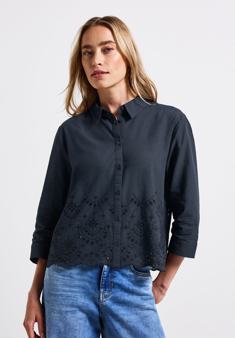Camicia nera con bottoni, orlo ricamato a scallop, maniche a tre quarti e motivi floreali testurizzati sul fondo. Sono esposti jeans in denim.
