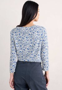 Langetermijn top in witte stof met een herhalend blauw cirkelpatroon. Kenmerkt zich door een ronde halslijn en een aansluitende silhoeette. Gecombineerd met donkere broeken.