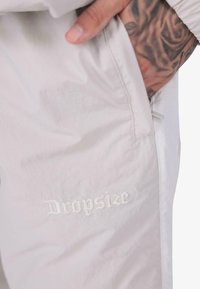 Tuvuma tuvuma ar roku ar tetovējumu, kas ievietota gaišo auduma biksu kabatā, uz kurām ir izšūts teksts "Dropsize".