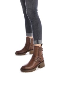 Stivali alla caviglia in pelle marrone con tacco spesso, dettagli con fibbia e cerniera laterale. Indossati con jeans scuri arrotolati. Suola robusta e strutturata.