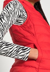 Rode gewatteerde vest met een hoge kraag, voorzien van een zijzak. Daaronder een zwart-wit gestreepte kleding met lange mouwen. Gestructureerde stof.