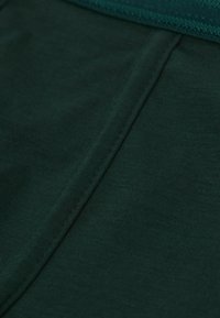Mörkgrön tyg med en slät textur, med en söm och ribbad midjeband. Materialet verkar vara elastiskt och lätt.