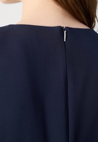 Gros plan sur le cou et le haut du dos d’une femme portant un vêtement bleu marine avec une petite tirette de fermeture éclair argentée au niveau de l’encolure arrière.