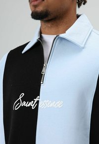Veste pour homme à demi-fermeture éclair, avec des blocs de couleur bleu et noir, un col à revers, et le logo "Saintessence" brodé en blanc. Matière en mélange de coton.