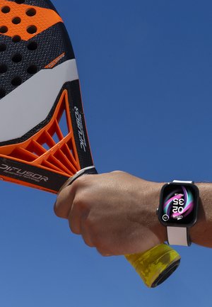 Hand gripping orange and black padel racket with yellow grip, wrist wearing smartwatch showing time and date, clear blue sky background.