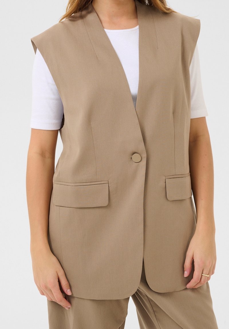 Blazer sans manches beige avec un bouton unique et des poches à rabat, porté sur une chemise blanche à manches courtes côtelée et un pantalon beige assorti.