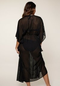 Kimono negro semitransparente con patrón geométrico, de frente abierto y cinturilla ajustable. Presenta mangas anchas y cae por debajo de la rodilla.