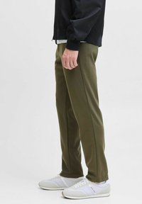 Pantalons de survêtement vert olive avec détails sur les coutures latérales, associés à des baskets blanches avec des accents gris et une veste bomber noire portée par-dessus.