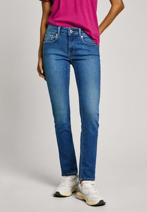 Vaqueros ajustados azules con un diseño clásico de cinco bolsillos, cierre de botón y un ligero desvanecimiento. Combinados con una camiseta rosa y zapatillas blancas.