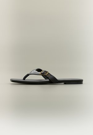 Sort flat sandal med tynn såle, tåthong og en sidestropp med detalj i messingspenne.