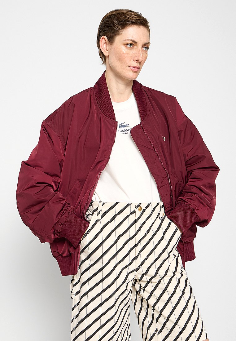 Veste bomber bordeaux avec des poignets et un col côtelés, présentant une texture lisse. Associée à un pantalon rayé crème et noir.