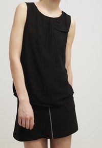 Blusa nera senza maniche con dettagli plissettati vicino al collo, abbinata a una minigonna nera con zip sul davanti.