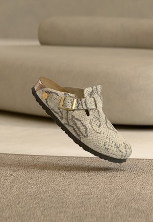 Zapato tipo slip-on con estampado de serpiente, hebilla dorada y suela negra, colocado sobre una alfombra frente a un sofá beige.