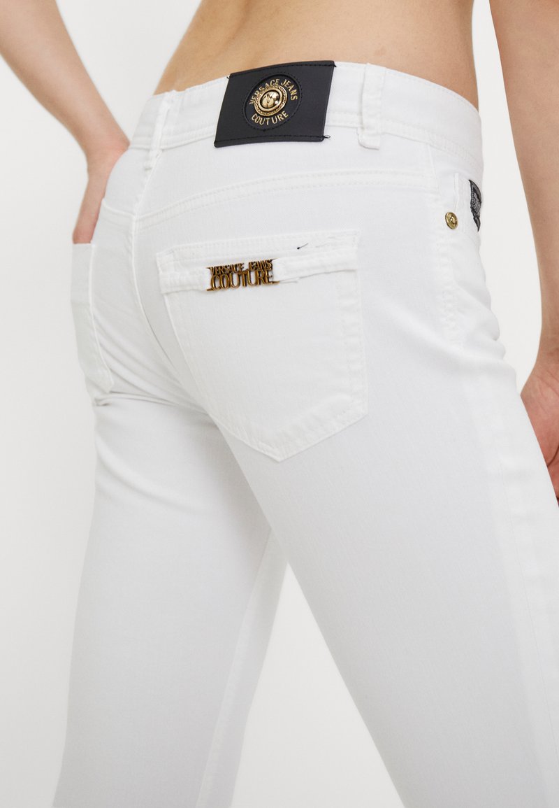 Witte denim jeans met een zwart label en een gouden logo aan de taille, en een gouden accent op de achterzak. Glad textuur.