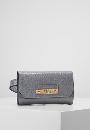 Grå clutch i imiteret krokodilleskind med lås i guldfarve og håndledsrem, placeret på en hvid piedestal mod en lysegrå baggrund.