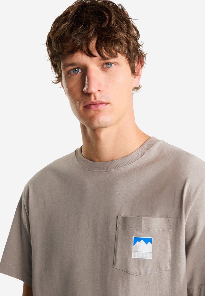 Joven con cabello castaño ondulado que lleva una camiseta color topo con un bolsillo en el pecho y un logo de montaña azul y blanco.