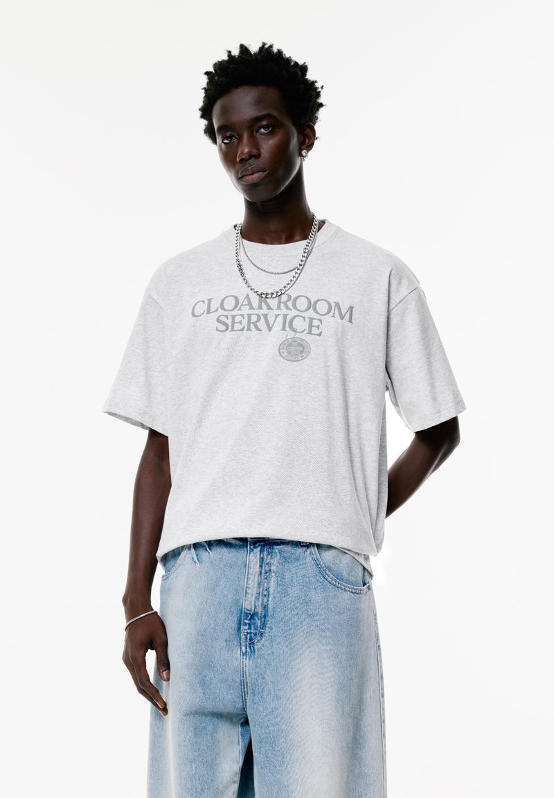 Tricou gri cu mânecă scurtă, având inscripția "Cloakroom Service" în gri închis, asortat cu blugi de denim bleumarin deschis. Design simplu, cu o croială lejeră.