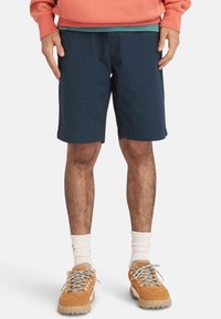 Donkerblauwe katoenen shorts met een ontspannen pasvorm, voorzien van zijzakken en een gestikte zoom, gecombineerd met tan sneakers en witte sokken.