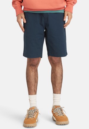Marinblå bomullsshorts med en avslappnad passform, som har sidofickor och söm i fållen, kombinerade med sandfärgade sneakers och vita strumpor.