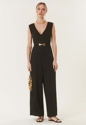 Femme portant une combinaison noire sans manches à col en V, ceinture dorée, sandales, et tenant une pochette à sequins dorés sur un fond uni.