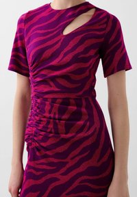 Aansluitende jurk in paarse stof met rode zebra strepen. Heeft korte mouwen en een uitsparing aan de zijkant. Gerimpelde details bij de taille.