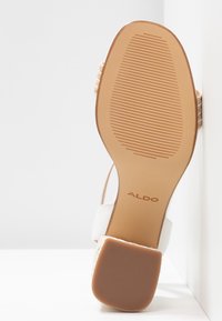 Sandale avec semelle en caoutchouc beige, petit talon texturé, lanières blanches et nom de la marque « ALDO » visible sur la semelle, positionnée à la verticale contre une surface blanche.