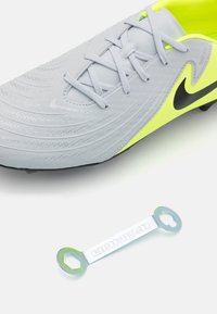 Grå syntetiske fodboldstøvler med en tekstureret overflade, sort Nike-logo og lysegule detaljer; inkluderer et metalværktøjslignende tilbehør nedenfor.