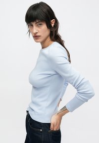 Jasnoniebieska, długorękawowa bluzka z prążkowanej tkaniny, z okrągłym dekoltem i dopasowanym krojem, zestawiona z ciemnymi dżinsami. Gładka faktura, minimalistyczne detale.